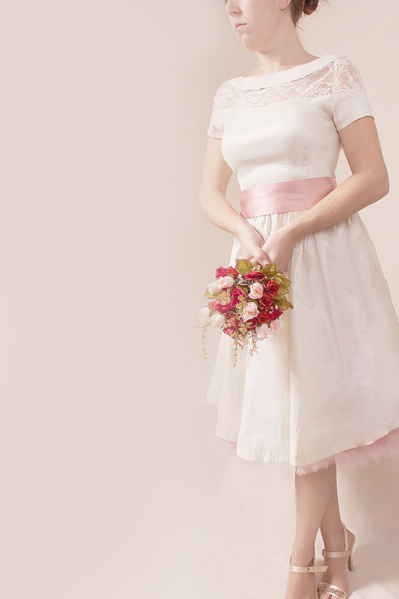 May include: A white dress with a lace bodice and a pink satin sash. The dress has a full skirt and is worn with a bouquet of pink and red roses.