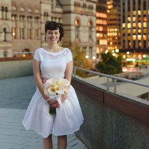 May include: A bride in a white dress with a lace bodice and a pink sash holds a bouquet of white calla lilies. She is standing on a sidewalk in front of a building.