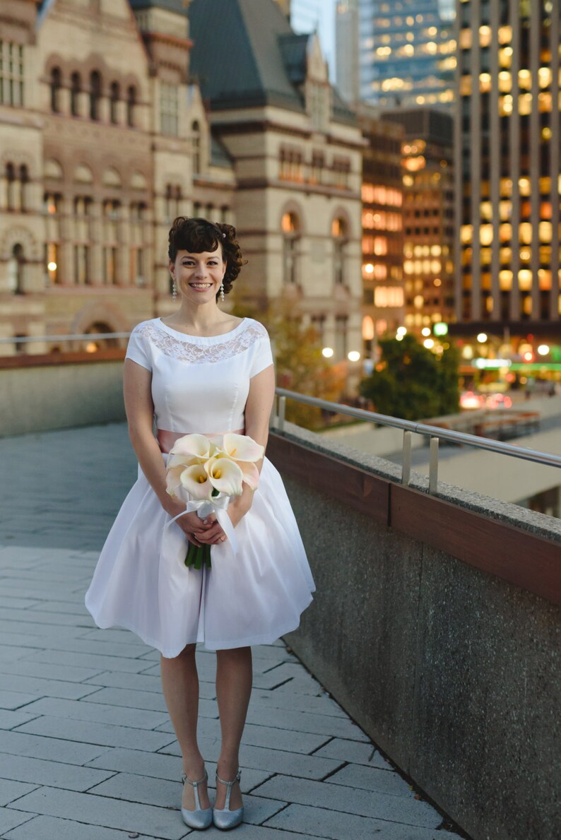 May include: A bride in a white dress with a lace bodice and a pink sash holds a bouquet of white calla lilies. She is standing on a sidewalk in front of a building.