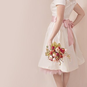 May include: A white dress with a pink sash and lace detailing. The dress has a tulle underskirt and the model is holding a bouquet of pink and red roses.