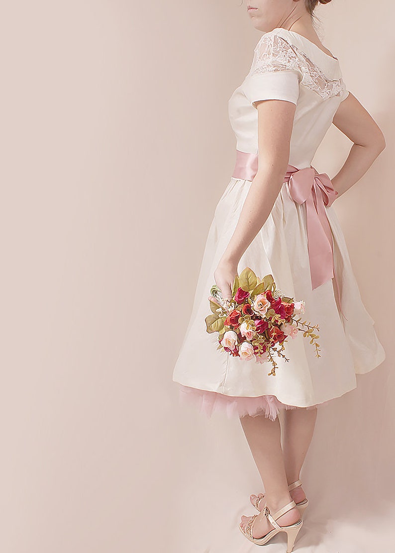 May include: A white dress with a pink sash and lace detailing. The dress has a tulle underskirt and the model is holding a bouquet of pink and red roses.