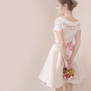 May include: A white dress with a lace bodice and a pink satin sash. The dress has a full skirt and is worn by a person holding a bouquet of red, pink, and green flowers.