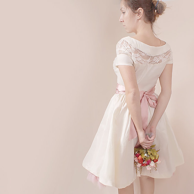 May include: A white dress with a lace bodice and a pink satin sash. The dress has a full skirt and is worn by a person holding a bouquet of red, pink, and green flowers.