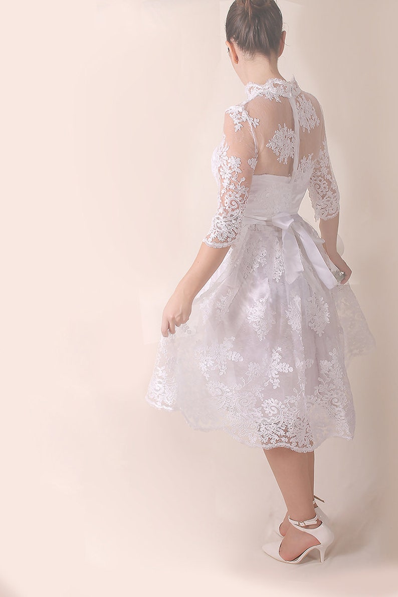 K&ouml;nnte beinhalten: Ein wei&szlig;es Spitzenkleid mit hohem Ausschnitt und einem taillierten Oberteil. Das Kleid hat einen vollen Rock und eine Satin-Sch&auml;rpe, die in der Taille gebunden ist. Das Kleid wird von einer Person getragen, die mit dem R&uuml;cken zur Kamera steht.