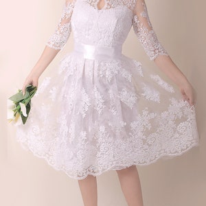 K&ouml;nnte beinhalten: Ein wei&szlig;es Spitzenkleid mit hohem Ausschnitt und einem figurbetonten Oberteil. Das Kleid hat einen vollen Rock und eine Satinschleife an der Taille. Das Kleid ist perfekt f&uuml;r eine Hochzeit oder einen anderen besonderen Anlass.