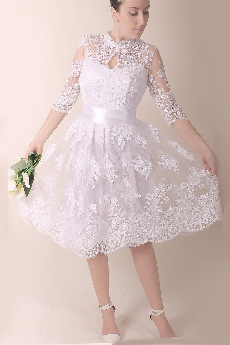K&ouml;nnte beinhalten: Ein wei&szlig;es Spitzenkleid mit hohem Ausschnitt und einem figurbetonten Oberteil. Das Kleid hat einen vollen Rock und eine Satinschleife an der Taille. Das Kleid ist perfekt f&uuml;r eine Hochzeit oder einen anderen besonderen Anlass.