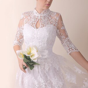 K&ouml;nnte beinhalten: Ein wei&szlig;es Spitzen-Hochzeitskleid mit hohem Ausschnitt, Dreiviertel&auml;rmeln und vollem Rock. Das Kleid hat eine Satinschleife in der Taille und ist mit zarten Blumenstickereien verziert.