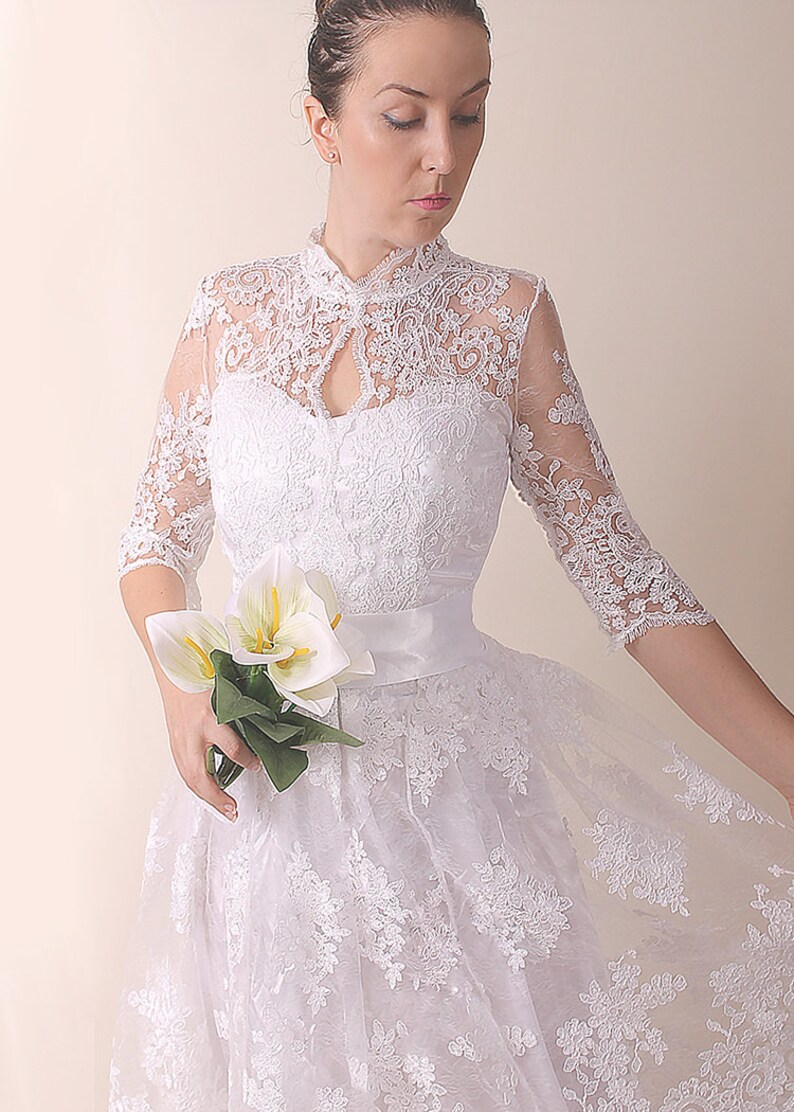 K&ouml;nnte beinhalten: Ein wei&szlig;es Spitzen-Hochzeitskleid mit hohem Ausschnitt, Dreiviertel&auml;rmeln und vollem Rock. Das Kleid hat eine Satinschleife in der Taille und ist mit zarten Blumenstickereien verziert.