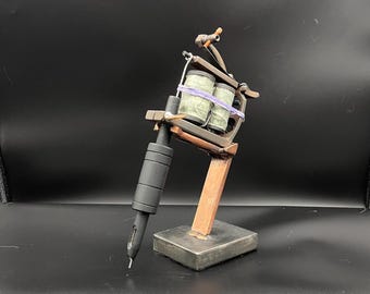 May include: A detailed model of a tattoo machine, featuring a black grip, needle, and a wooden frame. The machine has two spools wrapped with green material and secured with a purple band. It is mounted on a wooden stand with a square base.