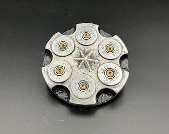 May include: A silver-colored, circular object resembling a revolver cylinder, featuring six Winchester 20 GA bullet casings. A star-shaped design is etched in the center. The object is set against a dark background.