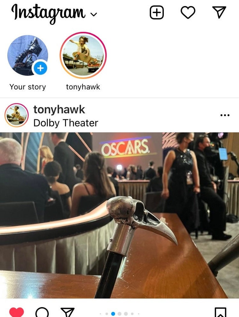 May include: A silver skull-shaped cane with a black handle. The cane is resting on a wooden table in front of a group of people sitting in chairs. The word "OSCARS" is visible in the background.