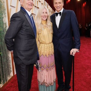 May include: Three people are standing on a red carpet. The person on the left is wearing a black suit, a blue scarf, and a black beret. The person in the middle is wearing a long, multi-colored dress with a yellow top and a pink, green, and blue bottom. The person on the right is wearing a dark blue suit, a white shirt, and a black bow tie. He is holding a black cane.