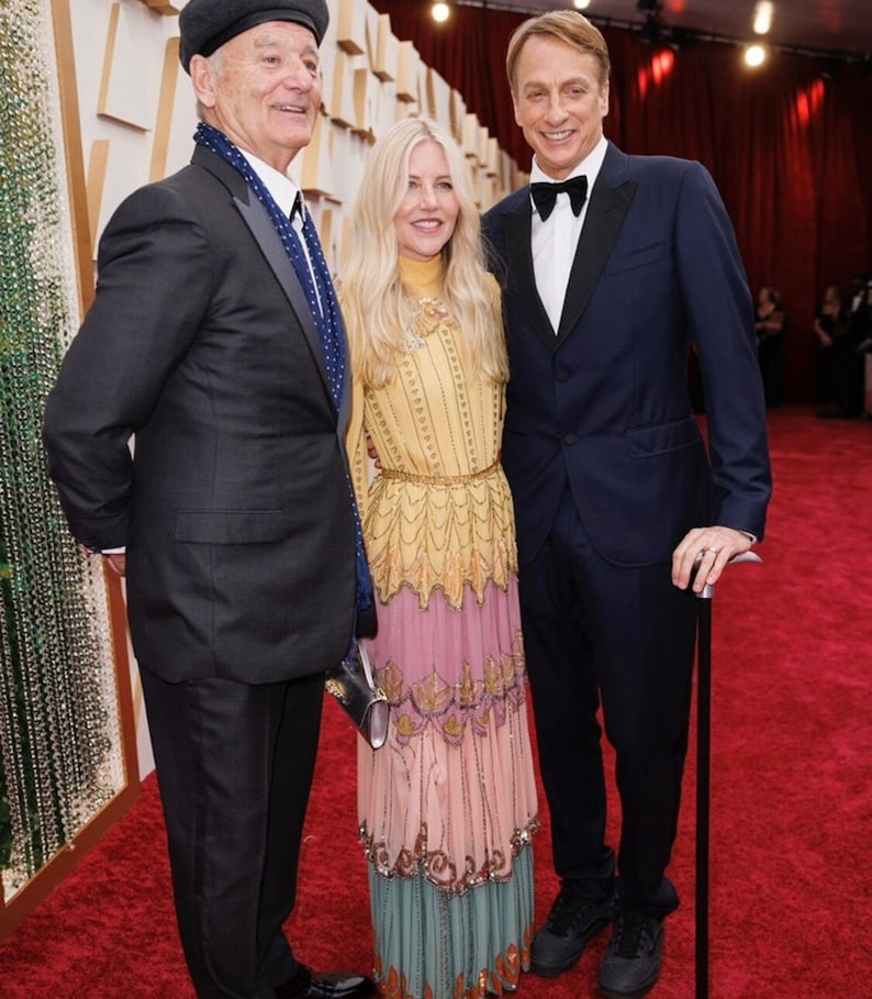 May include: Three people are standing on a red carpet. The person on the left is wearing a black suit, a blue scarf, and a black beret. The person in the middle is wearing a long, multi-colored dress with a yellow top and a pink, green, and blue bottom. The person on the right is wearing a dark blue suit, a white shirt, and a black bow tie. He is holding a black cane.