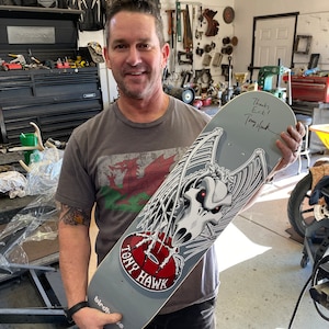 May include: A gray skateboard with a black and white graphic of a skull with wings. The skateboard is signed by Tony Hawk with the message "Thanks Erik!"