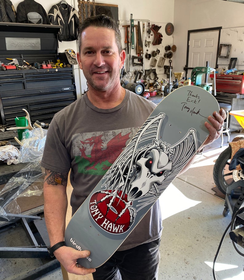 May include: A gray skateboard with a black and white graphic of a skull with wings. The skateboard is signed by Tony Hawk with the message "Thanks Erik!"