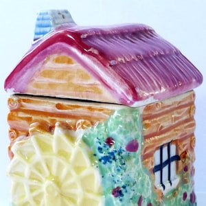 May include: A ceramic cottage-shaped sugar bowl with a red roof, brown log cabin walls, and a yellow windmill design. The bowl is decorated with green and pink floral details.