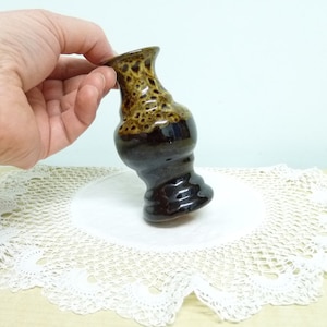 May include: A small brown and black ceramic vase with a speckled glaze. The vase is sitting on a white doily.