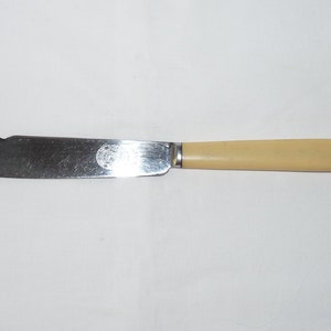 May include: A vintage butter knife with a stainless steel blade and a cream-colored handle. The blade has a curved edge and a decorative emblem near the handle. The knife is designed for spreading butter or other soft spreads.