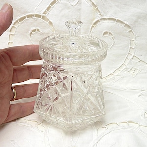 Puede incluir: Un tarro de vidrio transparente con un patrón de diamante y una tapa. El tarro está sobre un mantel de encaje blanco.