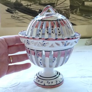 May include: A decorative ceramic lidded bowl with a pedestal base. The bowl features a white and pink striped design with floral accents. The lid has a handle and matches the bowl's design. The bowl is held by a hand.