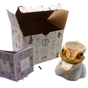 Könnte beinhalten: Eine weiße Keramiktasse in Form eines Frosches, der ein weißes Hemd trägt und ein Buch hält. Die Tasse befindet sich in einer Schachtel mit einem Buch und einem Bild eines Frosches auf der Schachtel.