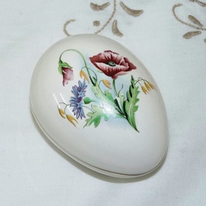 May include: White ceramic egg-shaped trinket box with a floral design. The design features a red poppy, blue and purple flowers, and green leaves.