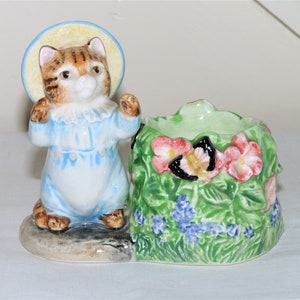 May include: A ceramic figurine of a cat wearing a blue and white outfit and a yellow hat, standing next to a green ceramic planter with pink flowers and a butterfly.