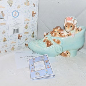 Puede incluir: Un zapato de cerámica azul con un grupo de figuritas de ratones en su interior. Los ratones son marrones y blancos con detalles rosas. El zapato está sobre una superficie blanca con una caja y un folleto en el fondo. La caja tiene un patrón azul y blanco con las palabras "El mundo de Beatrix Potter". El folleto tiene las palabras "El mundo de Beatrix Potter" en la portada.