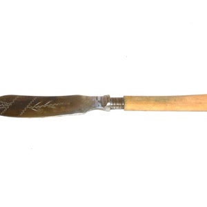 May include: A vintage butter knife with a light wooden handle and a silver blade. The blade features etched decorative patterns. The knife is designed for spreading butter or soft cheeses. The handle is a light tan color.