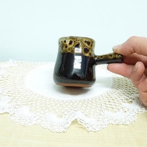 May include: A small, brown and black ceramic mug with a handle. The mug has a speckled glaze and is sitting on a white doily.