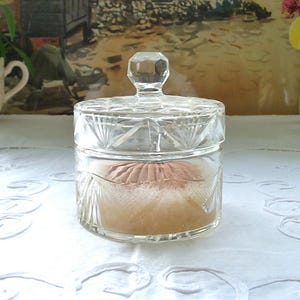 Könnte beinhalten: Ein klarer Puderbehälter aus Glas mit facettiertem Deckel und geätztem Design. Der Behälter enthält eine Puderquaste und steht auf einer weißen, bestickten Tischdecke. Eine Blumetasse und ein Gemälde sind im Hintergrund. Höhe ca. 10 cm.