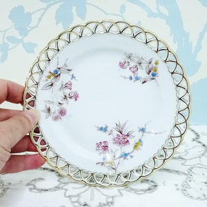 May include: A white porcelain plate with a gold rim and delicate floral pattern. The plate has a scalloped edge and a lace-like design around the rim.