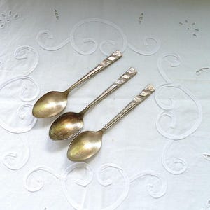 May include: Three antique silver spoons with patterned handles are displayed on a white, embroidered tablecloth. The spoons have oval bowls and detailed handles with diagonal lines. The spoons are arranged diagonally across the frame.