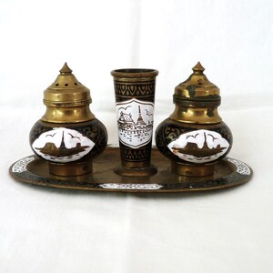 May include: A brass salt and pepper shaker set with a matching tray. The shakers have a black and gold design with white painted images of boats. The tray has a similar design.