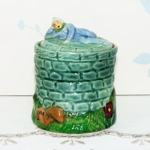 May include: A ceramic sugar jar with a green brick design and a lid featuring a figurine of a person lying on top. The jar has a green base with brown mushrooms and a red flower.