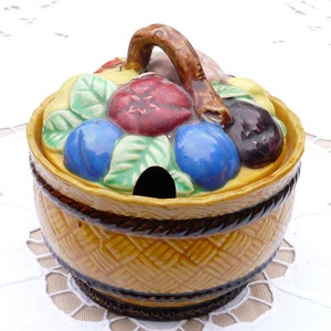 Pode incluir: Um açucareiro de cerâmica em forma de cesta com uma tampa decorada com frutas pintadas, incluindo ameixas, uvas e uma flor vermelha. A tampa tem uma alça marrom.