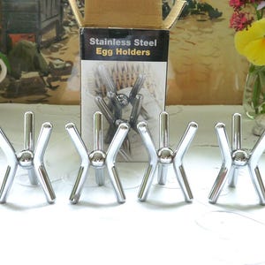 May include: Four stainless steel egg holders with a unique X-shaped design. The holders are displayed in front of a box that reads "Stainless Steel Egg Holders". The holders are on a white surface.