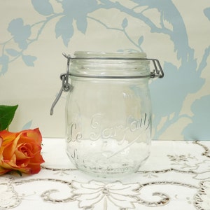 May include: A clear glass jar with a metal hinged lid and clasp. The jar has the words "Le Parfait" embossed on the front. An orange and yellow rose and green leaves are in the foreground.