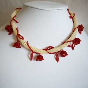 Peut inclure: Collier de perles crème avec des accents floraux rouges. Le collier est présenté sur un mannequin blanc. Les perles crème sont tissées en spirale, avec des fleurs et des feuilles rouges en perles.
