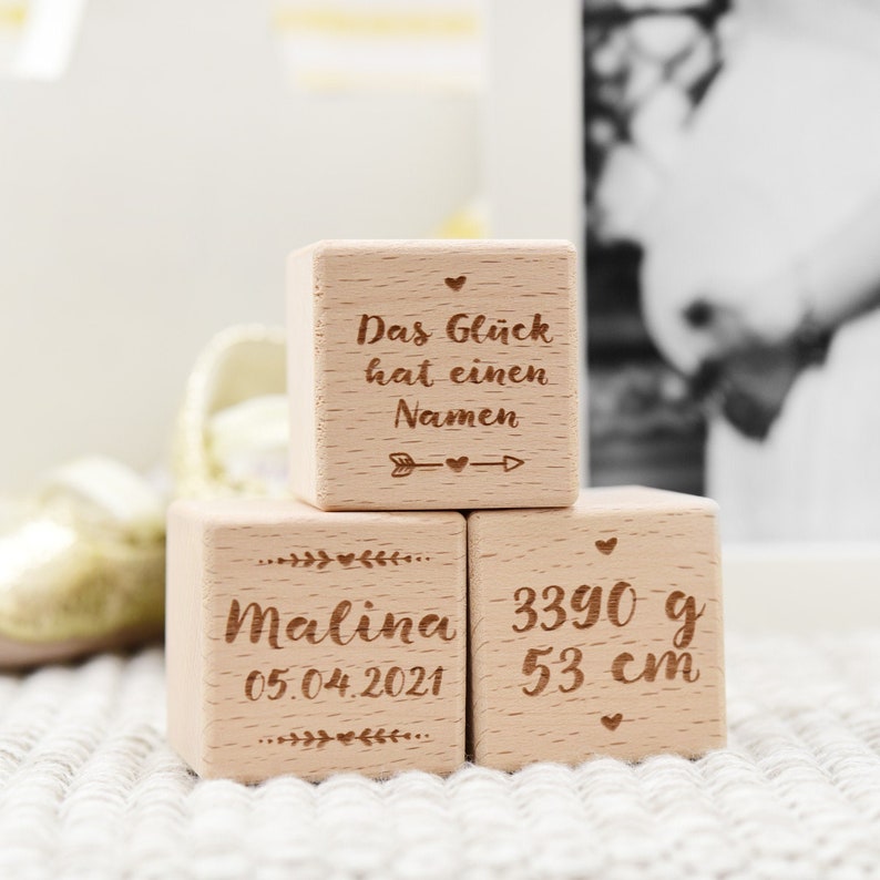 May include: Three wooden blocks with engraved text. The top block reads "Das Gl&uuml;ck hat einen Namen" with an arrow pointing to the right. The middle block reads "Malina 05.04.2021". The bottom block reads "3390 g 53 cm".