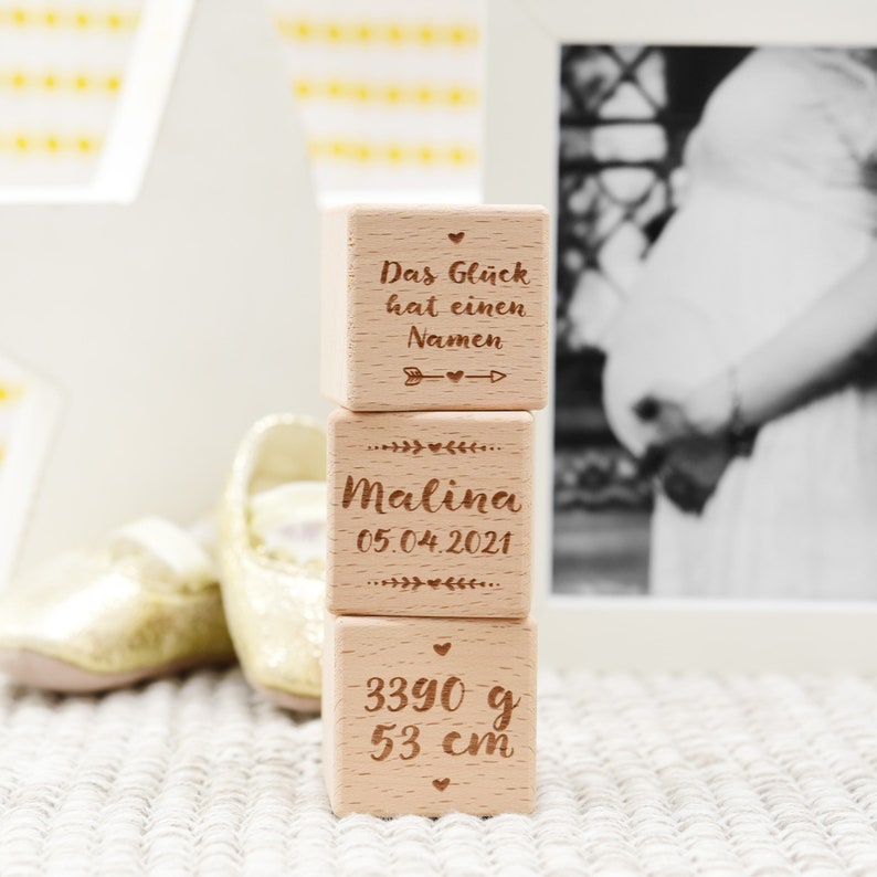 May include: Three wooden blocks stacked on top of each other. The top block reads "Das Gl&uuml;ck hat einen Namen" with an arrow pointing to the right. The middle block reads "Malina 05.04.2021". The bottom block reads "3390 g 53 cm".