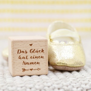 May include: A wooden block with the text "Das Gl&uuml;ck hat einen Namen" (German for "Happiness has a name") engraved on it. The block is sitting on a white knitted surface next to a gold baby shoe.