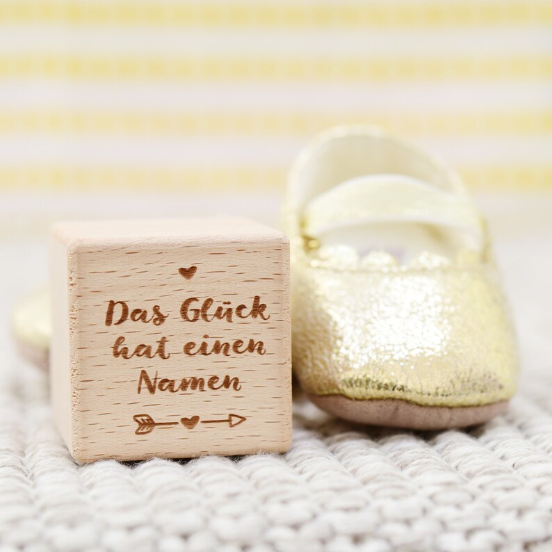 May include: A wooden block with the text "Das Gl&uuml;ck hat einen Namen" (German for "Happiness has a name") engraved on it. The block is sitting on a white knitted surface next to a gold baby shoe.