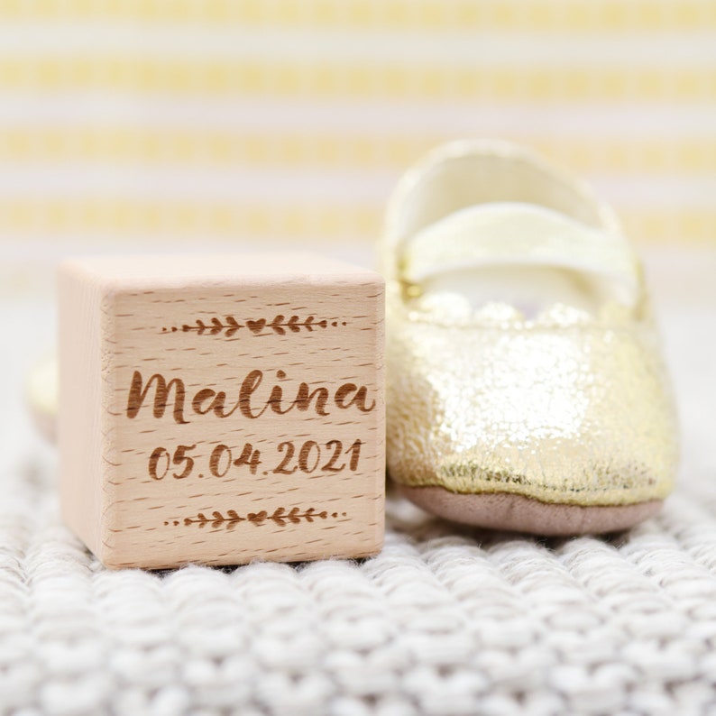 May include: A wooden block with the name Malina and the date 05.04.2021 engraved on it. A gold baby shoe is next to the block.
