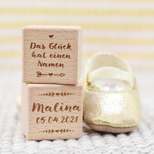 May include: Two wooden blocks stacked on top of each other. The top block has the text "Das Gl&uuml;ck hat einen Namen" with a small heart and arrow. The bottom block has the text "Malina 05.04.2021" with small hearts and leaves.
