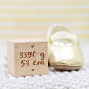 May include: A wooden block with the inscription "3390 g 53 cm" and two small hearts on either side. A gold baby shoe is next to the block.