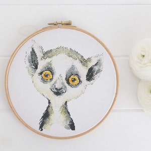 May include: A cross-stitch embroidery of a lemur's face, set in a wooden hoop. The lemur features large yellow eyes, grey and white fur, and a small black nose. The background is white.