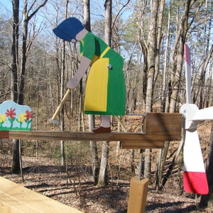 May include: A wooden folk art whirligig featuring a woman in a green and yellow dress, blue bonnet, and brown shoes, working with a hoe. The whirligig has a flower garden and a windmill with red and white blades. The background is a forest.