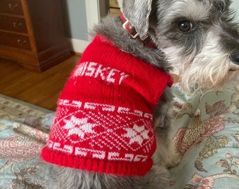May include: A small, gray dog wearing a red and white knitted sweater with the word "MISKEY" on the front. The sweater has a snowflake pattern.