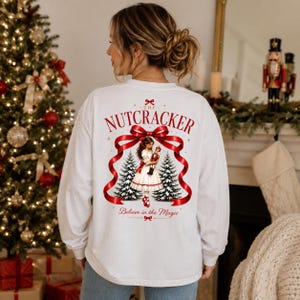 May include: A white long-sleeve shirt featuring a graphic on the back with the text THE NUTCRACKER and Believe in the Magic. The design displays a girl in a white dress holding a nutcracker, framed by a red ribbon and two pine trees.